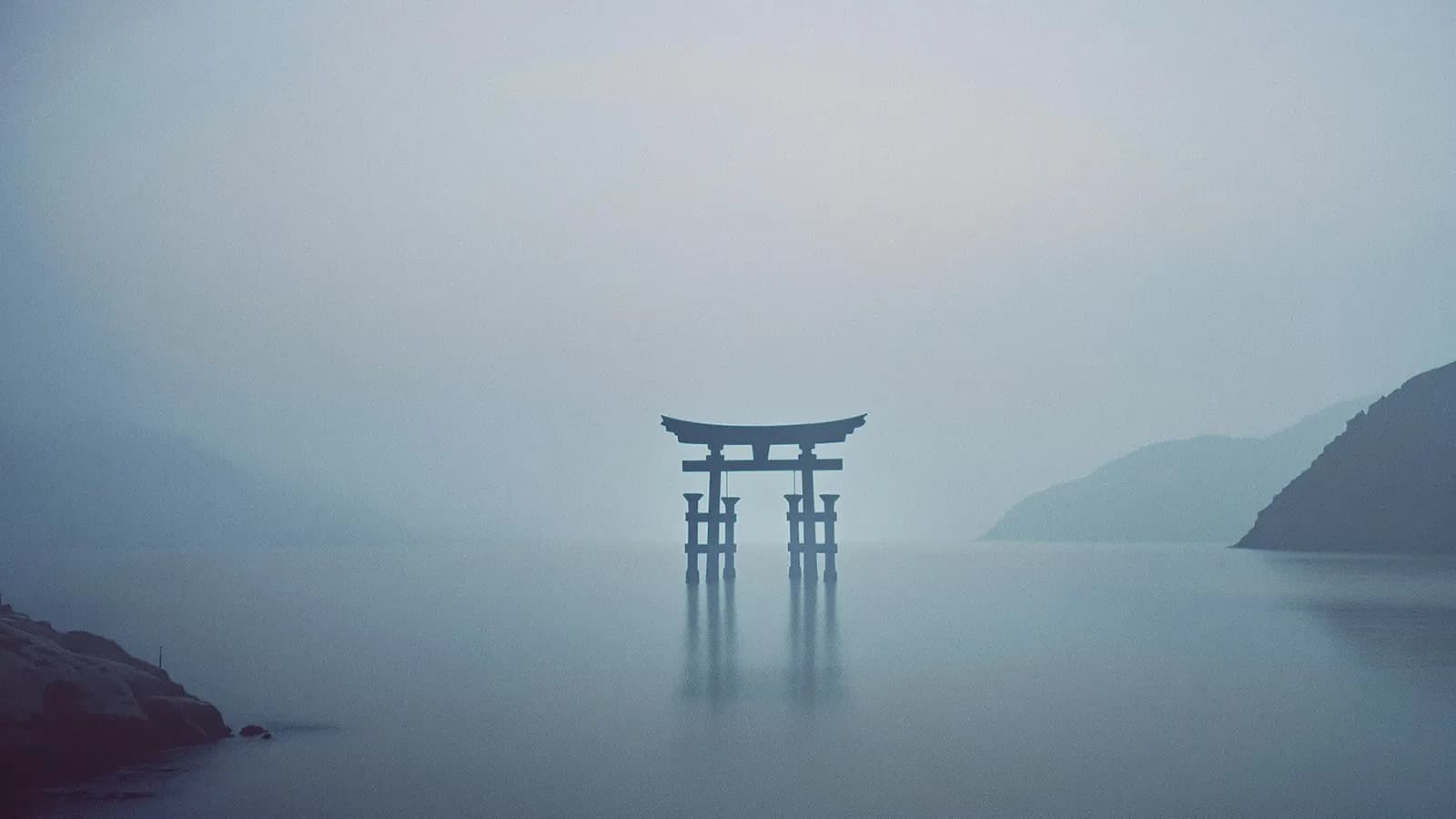A single torii gate standing in calm misty water at dawn, hazy hills behind