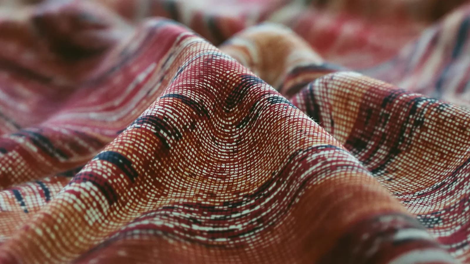 Extreme close-up of folded hand-woven ikat silk, bands of crimson, indigo and ochre blurring along the weave