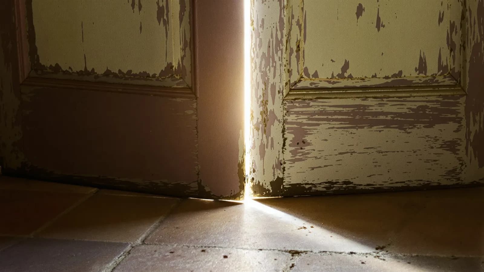 A narrow gap where an old painted wooden door stands slightly ajar, daylight spilling onto a worn stone floor